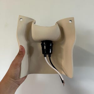 May include: A beige ceramic wall sconce fixture with a curved, molded shape featuring two circular mounting holes at the top corners. A black plastic light socket is centered in the middle, with one white and one black electrical wire extending.