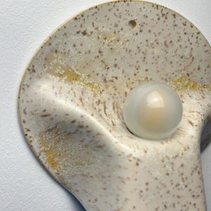 May include: A wall-mounted ceramic light fitting with a curved, shell-like shape. The surface features a speckled cream and brown glaze with yellow accents. A single, round, frosted glass light bulb is centred within the concave form.