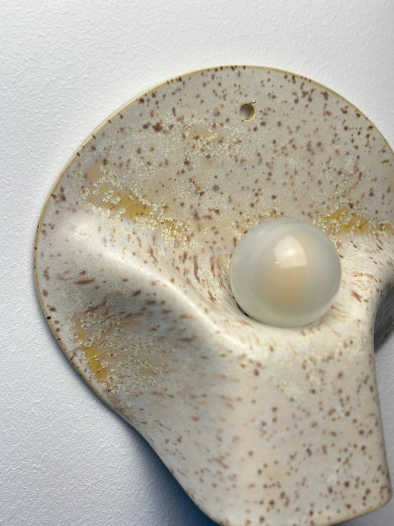 May include: A wall-mounted ceramic light fitting with a curved, shell-like shape. The surface features a speckled cream and brown glaze with yellow accents. A single, round, frosted glass light bulb is centred within the concave form.