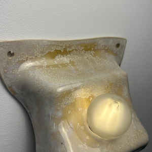 May include: A wall-mounted light fixture featuring a curved, off-white ceramic shade with a crystalline, crackled glaze finish in shades of amber and cream. The fixture includes a single, round, frosted glass light bulb centered on the shade.
