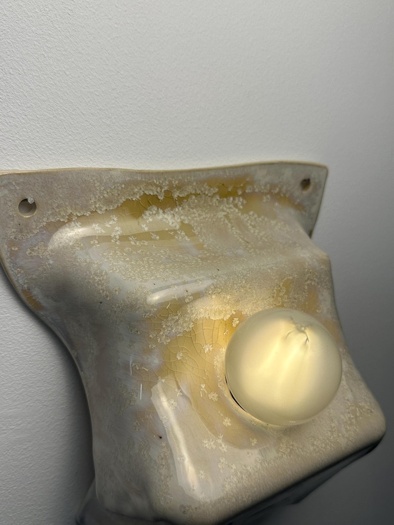 May include: A wall-mounted light fixture featuring a curved, off-white ceramic shade with a crystalline, crackled glaze finish in shades of amber and cream. The fixture includes a single, round, frosted glass light bulb centered on the shade.