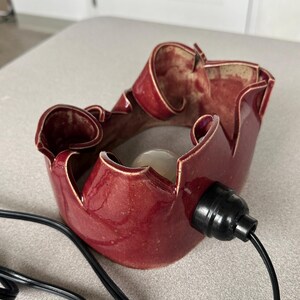 May include: A handcrafted, burgundy-colored ceramic lamp with a unique, ruffled design. The lamp features a black electrical socket and cord. The interior of the lamp is a lighter shade of red. The lamp is designed to hold a light bulb.