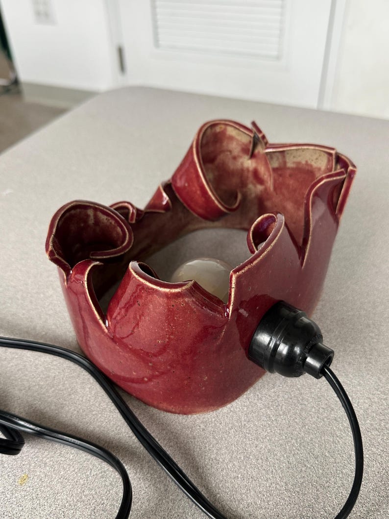May include: A handcrafted, burgundy-colored ceramic lamp with a unique, ruffled design. The lamp features a black electrical socket and cord. The interior of the lamp is a lighter shade of red. The lamp is designed to hold a light bulb.