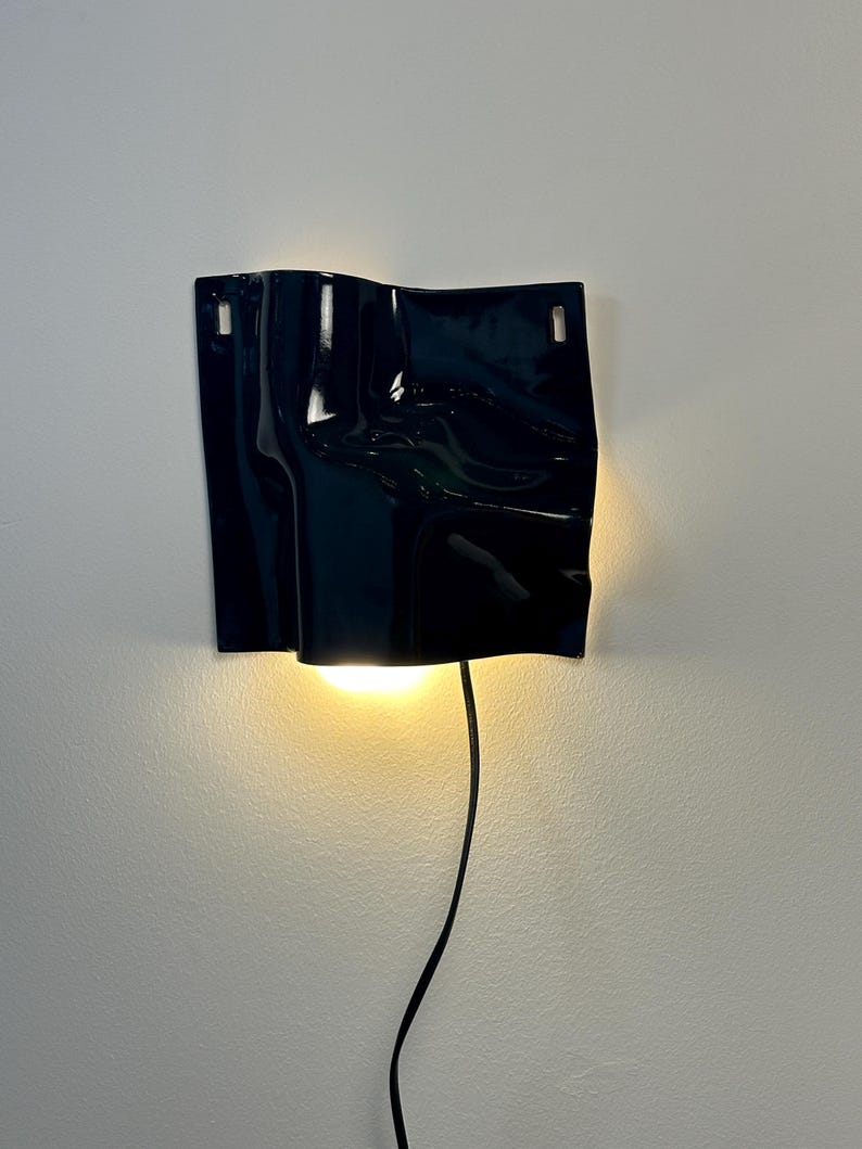 Puede incluir: Un aplique de pared negro brillante con una textura de superficie ondulada y plegada. La l&aacute;mpara cuenta con dos orificios de montaje rectangulares en la parte superior y emite una luz c&aacute;lida hacia abajo. Un cable negro sale de la base.
