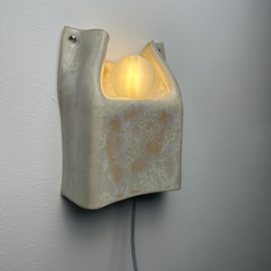 May include: A wall-mounted ceramic wall light with a cream-coloured, textured glaze. The fixture features a U-shaped opening at the top that houses a single, glowing, frosted glass spherical light bulb. A thin grey power cord extends from the bottom.