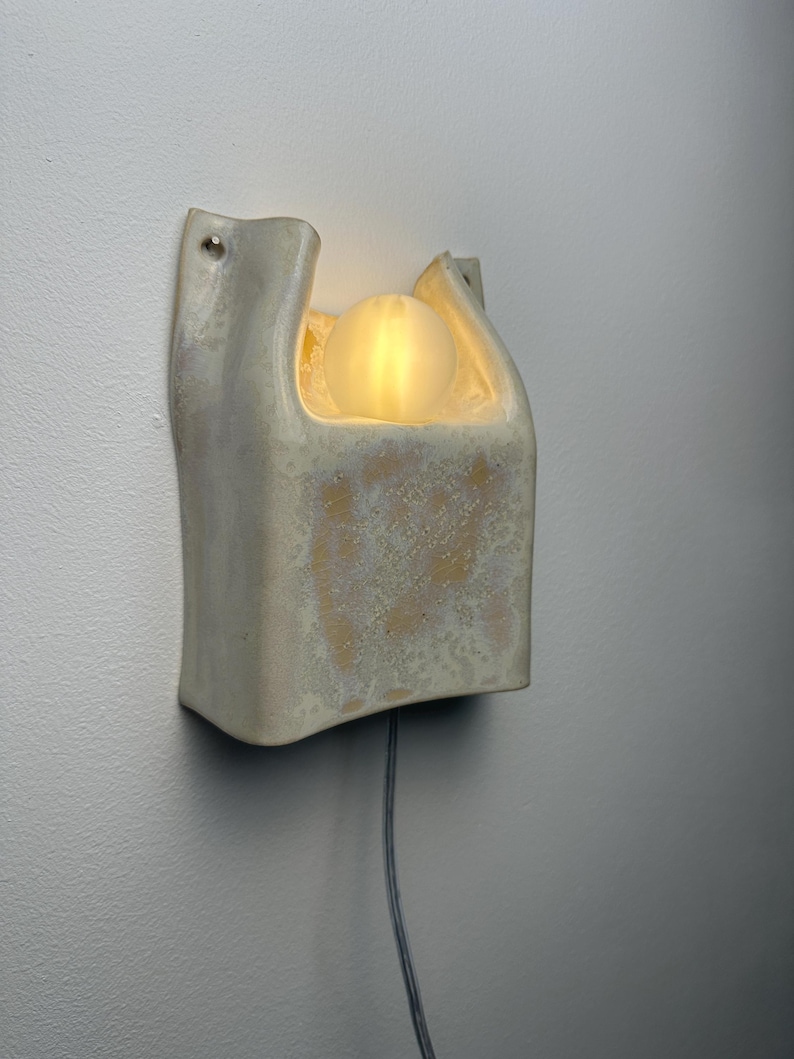 May include: A wall-mounted ceramic wall light with a cream-coloured, textured glaze. The fixture features a U-shaped opening at the top that houses a single, glowing, frosted glass spherical light bulb. A thin grey power cord extends from the bottom.
