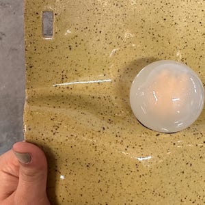 May include: A rectangular ceramic wall piece with a glossy mustard yellow glaze and dark brown speckles. The surface features a smooth, translucent white glass orb attached to the right side and a small rectangular hole at the top left corner.