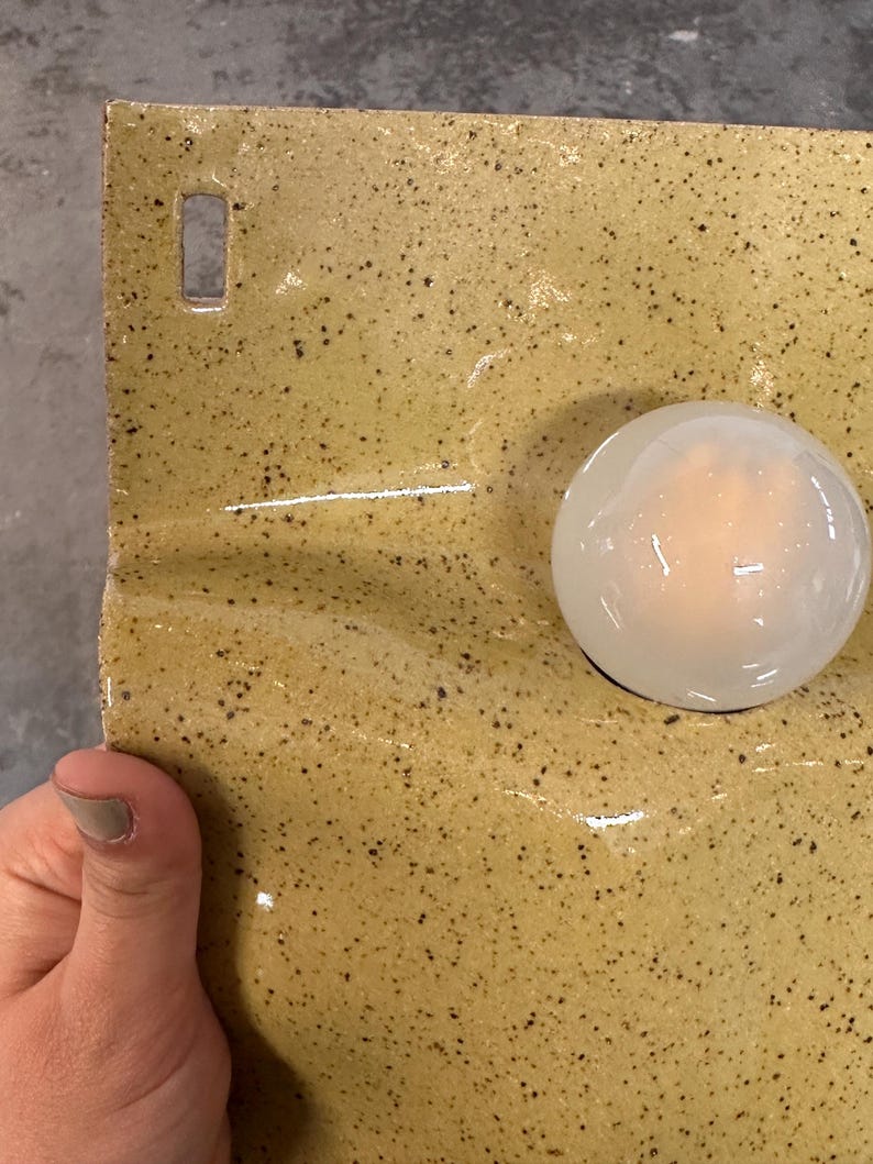 May include: A rectangular ceramic wall piece with a glossy mustard yellow glaze and dark brown speckles. The surface features a smooth, translucent white glass orb attached to the right side and a small rectangular hole at the top left corner.