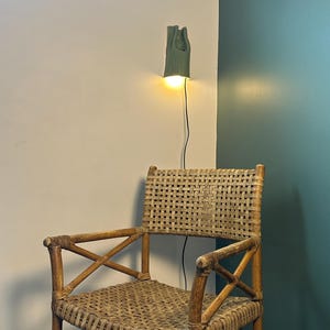 May include: A woven rattan armchair with a high backrest and cross-braced armrests. Above the chair, a sage green wall-mounted lamp emits a warm, glowing light, with a thin black power cord extending downward from the fixture to the floor.