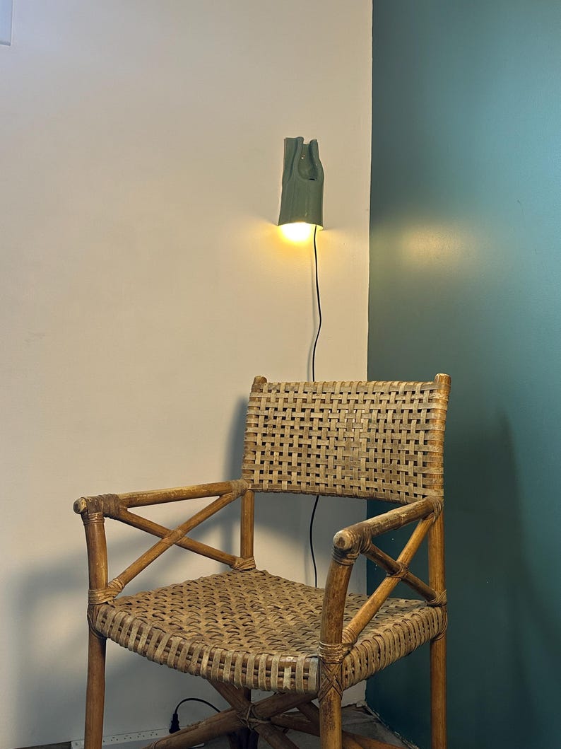 May include: A woven rattan armchair with a high backrest and cross-braced armrests. Above the chair, a sage green wall-mounted lamp emits a warm, glowing light, with a thin black power cord extending downward from the fixture to the floor.