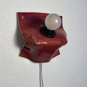 Puede incluir: Un aplique de pared con una base de cer&aacute;mica roja oscura brillante con una textura plegada similar a la tela. La l&aacute;mpara incluye un casquillo circular negro que sostiene una bombilla blanca esmerilada y un cable el&eacute;ctrico fino y transparente.