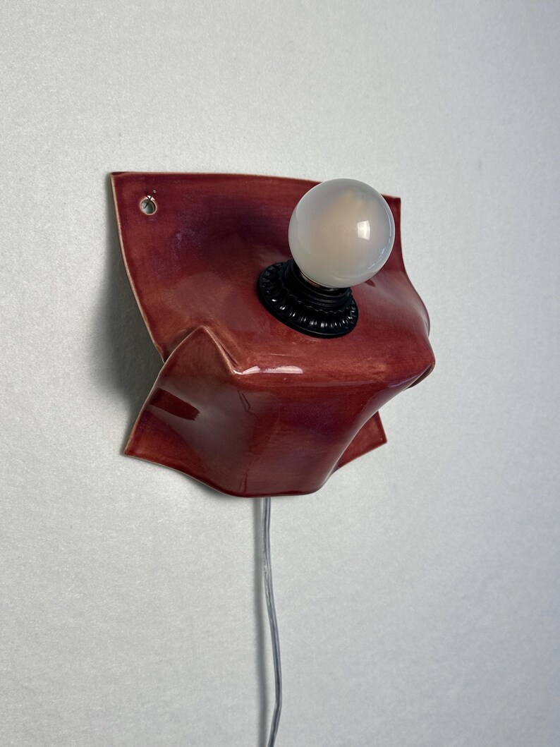 Puede incluir: Un aplique de pared con una base de cer&aacute;mica roja oscura brillante con una textura plegada similar a la tela. La l&aacute;mpara incluye un casquillo circular negro que sostiene una bombilla blanca esmerilada y un cable el&eacute;ctrico fino y transparente.