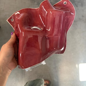 Puede incluir: Un bolsillo de pared de cer&aacute;mica de color rojo intenso brillante con una forma org&aacute;nica irregular. La pieza presenta una superficie lisa y reflectante y dos peque&ntilde;os orificios circulares en las esquinas superiores para su montaje. Un peque&ntilde;o lazo.