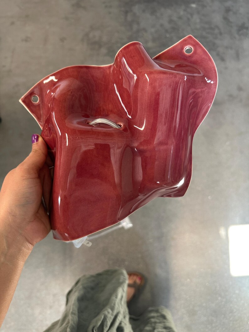 Puede incluir: Un bolsillo de pared de cer&aacute;mica de color rojo intenso brillante con una forma org&aacute;nica irregular. La pieza presenta una superficie lisa y reflectante y dos peque&ntilde;os orificios circulares en las esquinas superiores para su montaje. Un peque&ntilde;o lazo.
