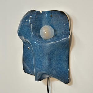 May include: A wall-mounted ceramic light fixture with a glossy, mottled blue glaze. The organic, undulating form features a central circular white glass bulb. A black power cord extends from the bottom of the piece, which includes two small mounting holes.