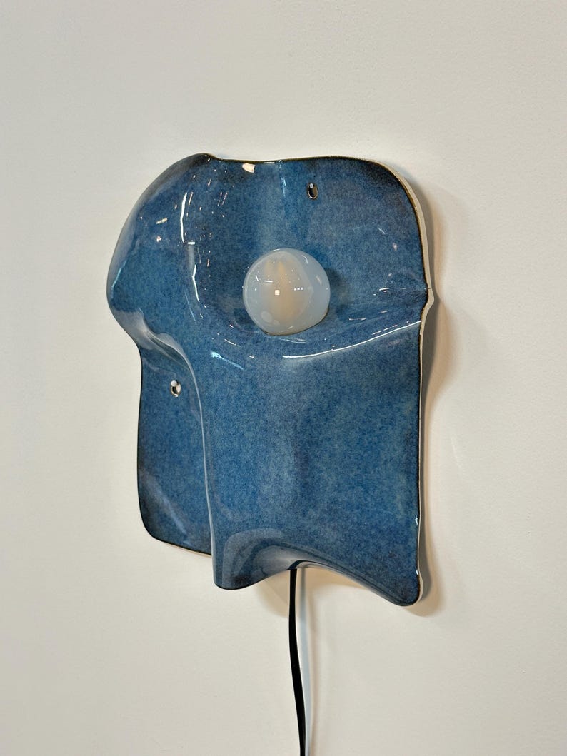 May include: A wall-mounted ceramic light fixture with a glossy, mottled blue glaze. The organic, undulating form features a central circular white glass bulb. A black power cord extends from the bottom of the piece, which includes two small mounting holes.