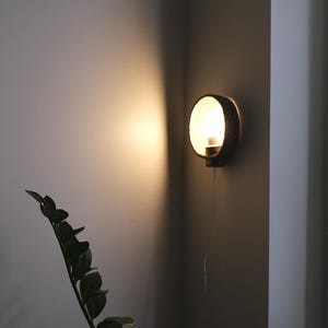 May include: A round, dark-colored wall sconce with a bright light emanating from within. The sconce is mounted on a corner wall, casting a warm glow. A plant with dark green leaves is in the foreground.