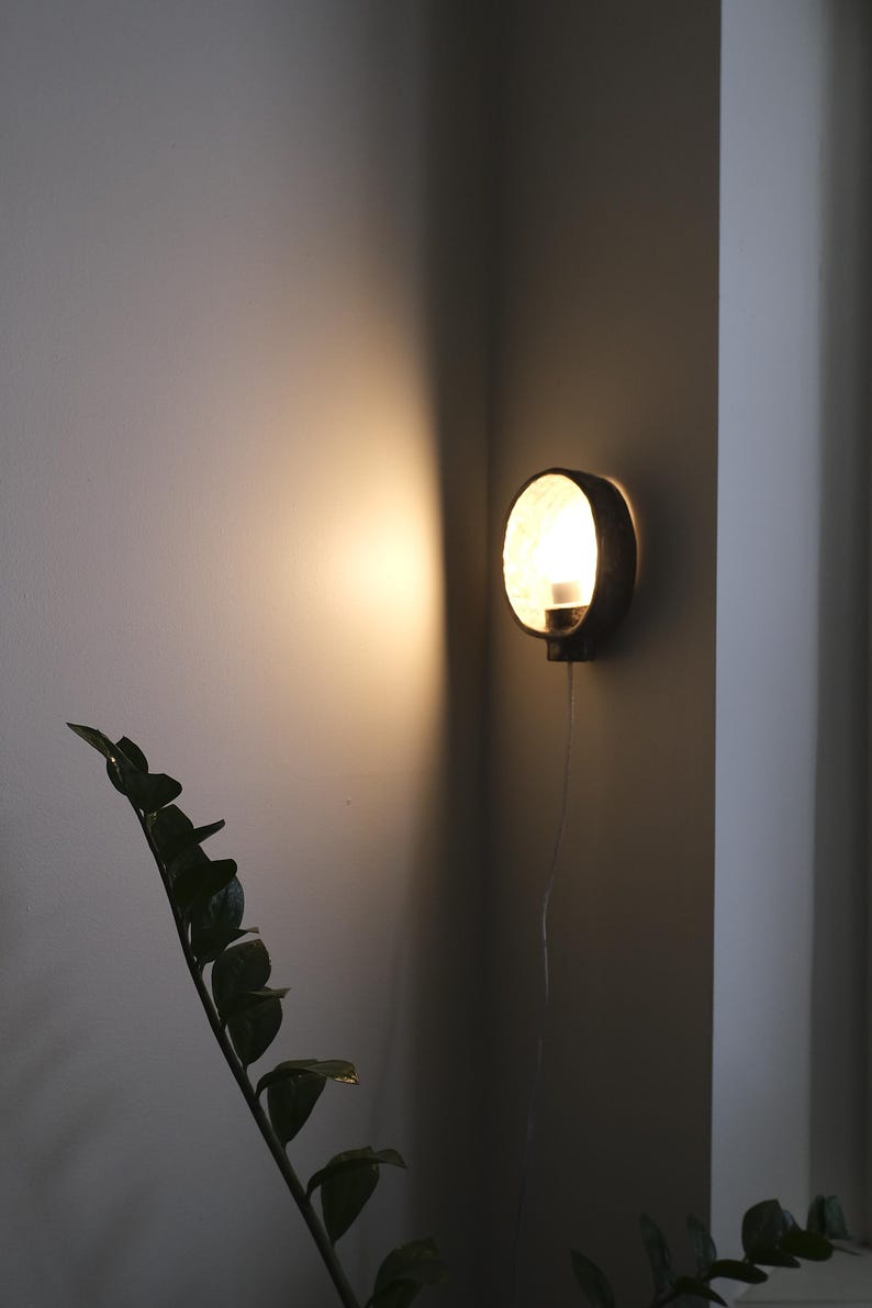 May include: A round, dark-colored wall sconce with a bright light emanating from within. The sconce is mounted on a corner wall, casting a warm glow. A plant with dark green leaves is in the foreground.
