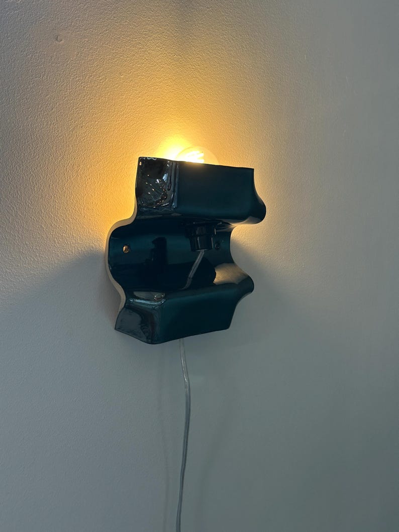 May include: A dark teal ceramic wall sconce with a glossy finish and an undulating, organic shape. The fixture features a central light bulb socket emitting a warm glow, with a clear power cord extending downward from the base of the unit.