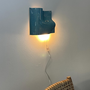 May include: A teal ceramic wall sconce with a speckled finish and a curved, sculptural form. The fixture emits a warm, downward-facing light and includes a thin, transparent power cord with an inline switch hanging down the wall.