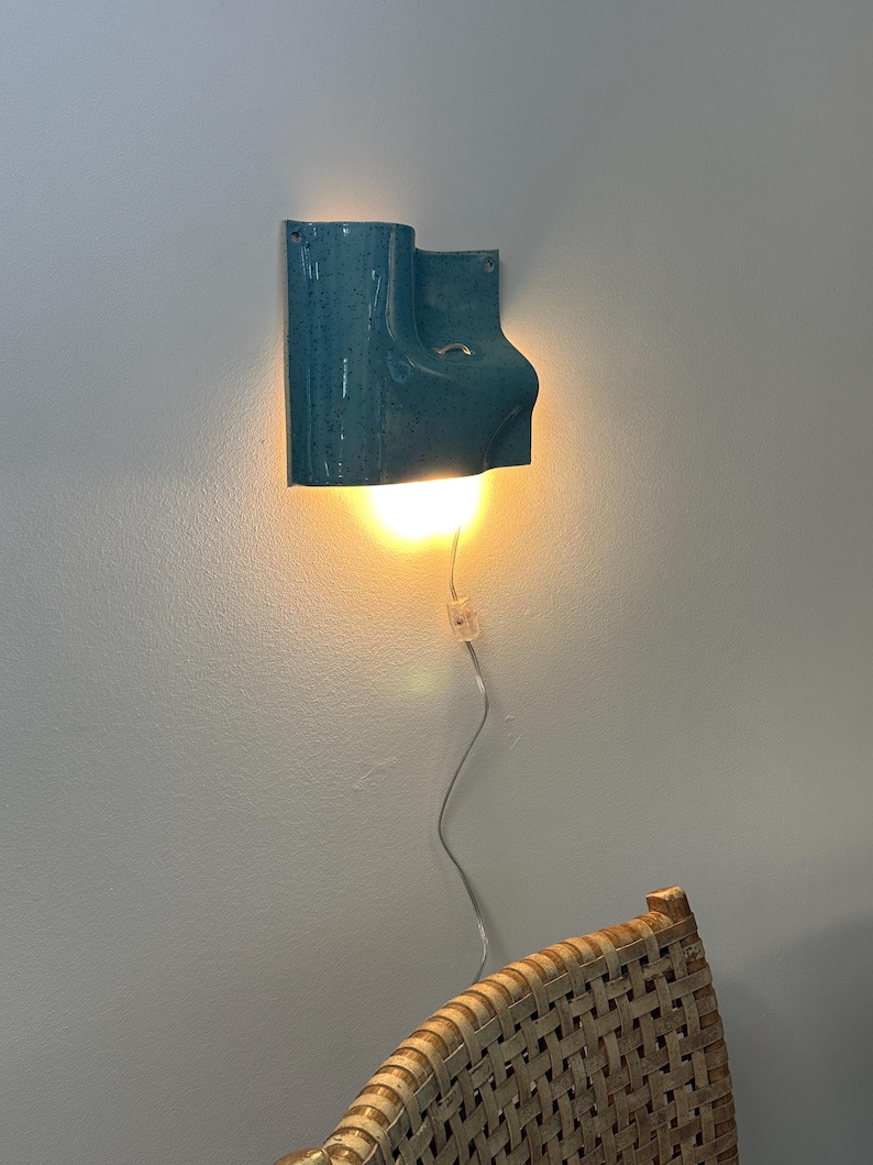 May include: A teal ceramic wall sconce with a speckled finish and a curved, sculptural form. The fixture emits a warm, downward-facing light and includes a thin, transparent power cord with an inline switch hanging down the wall.