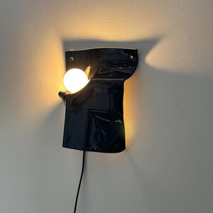 Puede incluir: Un aplique de pared con un escudo de cer&aacute;mica o metal negro brillante de forma irregular y superficie lisa y ondulada. Una bombilla blanca redonda e iluminada se sit&uacute;a en un hueco en el lado izquierdo, emitiendo una luz c&aacute;lida.