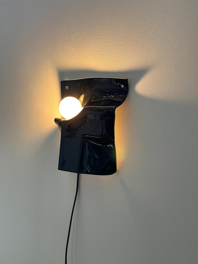 Puede incluir: Un aplique de pared con un escudo de cer&aacute;mica o metal negro brillante de forma irregular y superficie lisa y ondulada. Una bombilla blanca redonda e iluminada se sit&uacute;a en un hueco en el lado izquierdo, emitiendo una luz c&aacute;lida.