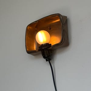 May include: A wall-mounted light fixture with a gold-colored interior and a matte bronze exterior. The fixture houses a round, illuminated light bulb. A black cord extends from the base of the fixture.