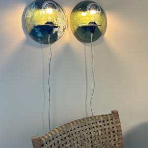May include: Two circular wall-mounted ceramic sconces with a glossy green glaze. Each fixture features a black socket holding a clear glass light bulb that emits a warm glow. A thin white power cord hangs vertically from each light fixture.