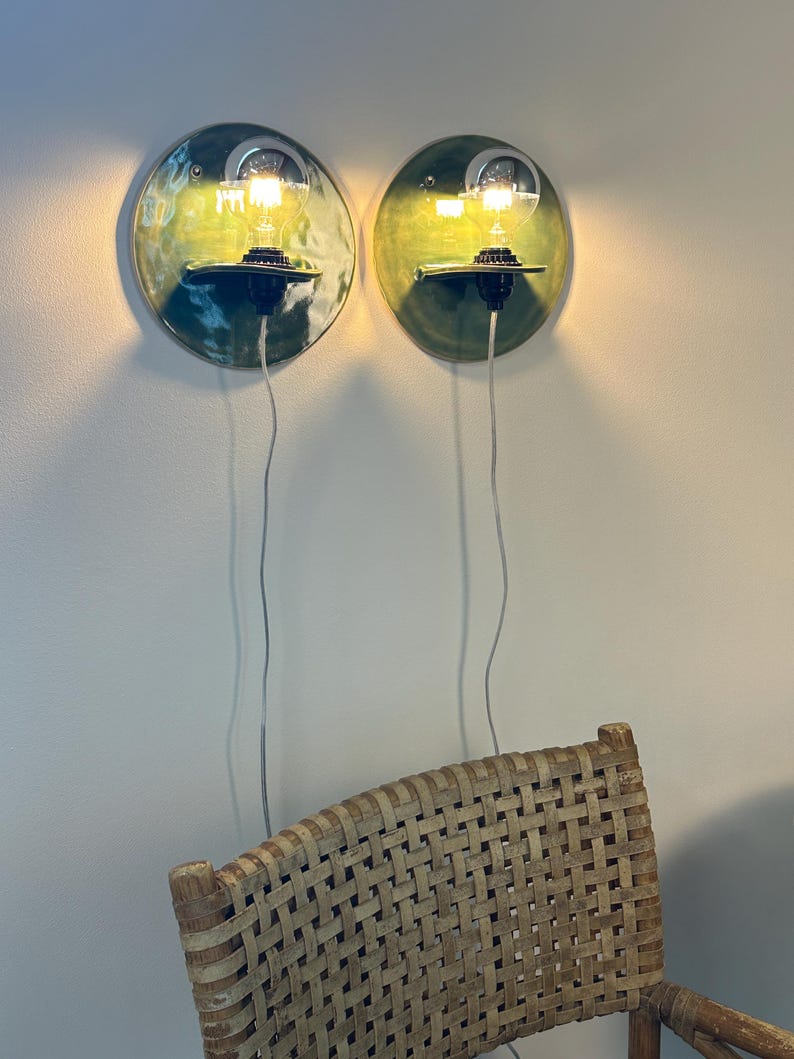 May include: Two circular wall-mounted ceramic sconces with a glossy green glaze. Each fixture features a black socket holding a clear glass light bulb that emits a warm glow. A thin white power cord hangs vertically from each light fixture.