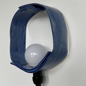 Puede incluir: Aplique de pared de cer&aacute;mica azul con un dise&ntilde;o curvo y abierto. El aplique contiene una bombilla blanca y un cable negro. La l&aacute;mpara est&aacute; montada en una pared blanca.
