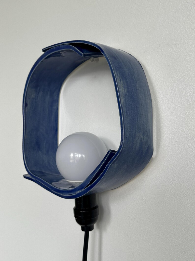 Puede incluir: Aplique de pared de cer&aacute;mica azul con un dise&ntilde;o curvo y abierto. El aplique contiene una bombilla blanca y un cable negro. La l&aacute;mpara est&aacute; montada en una pared blanca.