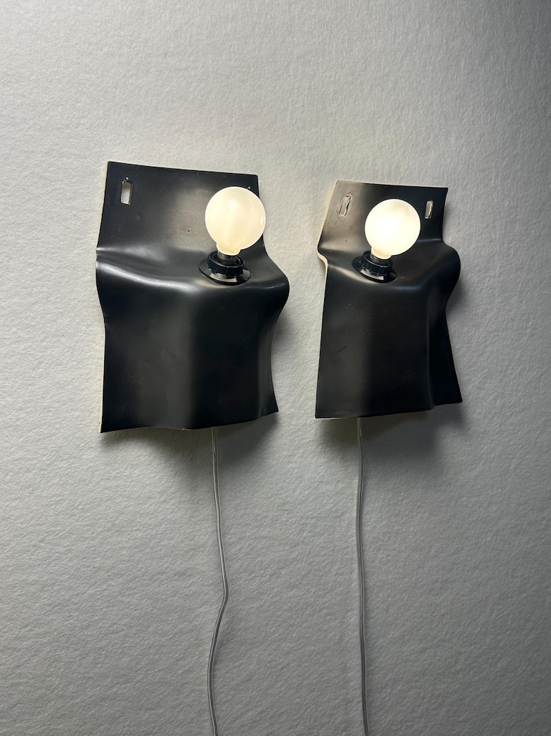 May include: A pair of wall-mounted lamps featuring black, flexible, rectangular panels that curve outward. Each lamp has a single white, round light bulb protruding from the center and a thin, light-colored electrical cord extending downward.