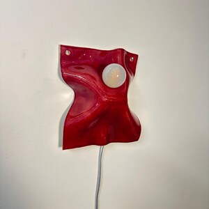 Puede incluir: Una l&aacute;mpara de pared con una base de cer&aacute;mica o resina de color rojo intenso, brillante y ondulada, con una forma fluida y org&aacute;nica. Una bombilla redonda de color blanco esmerilado est&aacute; centrada en la superficie. Un cable el&eacute;ctrico fino cuelga.