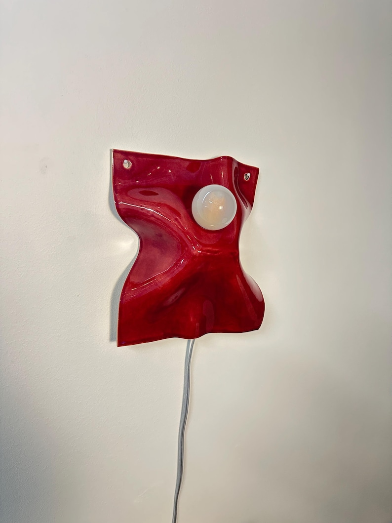 Puede incluir: Una l&aacute;mpara de pared con una base de cer&aacute;mica o resina de color rojo intenso, brillante y ondulada, con una forma fluida y org&aacute;nica. Una bombilla redonda de color blanco esmerilado est&aacute; centrada en la superficie. Un cable el&eacute;ctrico fino cuelga.