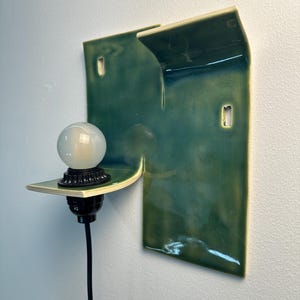 May include: A wall-mounted light fixture featuring a glossy, deep green ceramic backplate with an irregular, folded shape. A black socket holds a round, frosted white glass bulb, and a black power cord extends downward from the base of the socket.