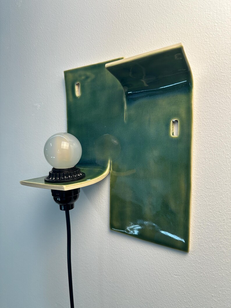 May include: A wall-mounted light fixture featuring a glossy, deep green ceramic backplate with an irregular, folded shape. A black socket holds a round, frosted white glass bulb, and a black power cord extends downward from the base of the socket.