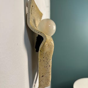 May include: A wall-mounted light fixture featuring a curved, cream-colored ceramic plate with a speckled brown pattern. The fixture includes a black socket holding a round, frosted white glass light bulb and a white electrical cord extending downward.