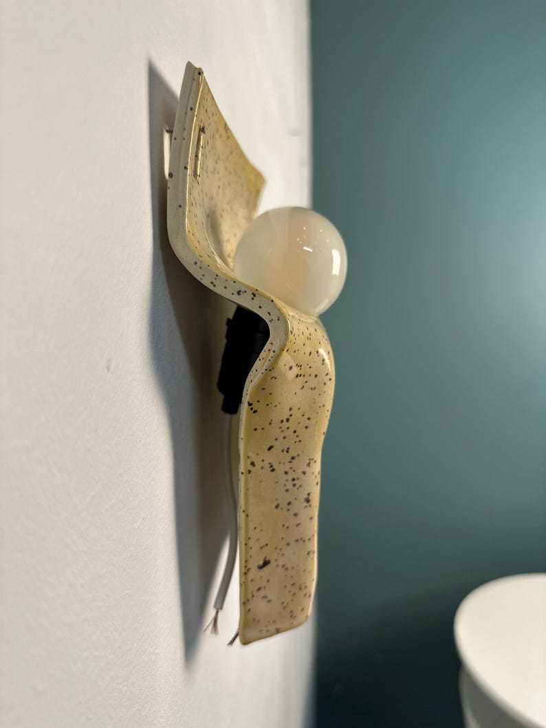 May include: A wall-mounted light fixture featuring a curved, cream-colored ceramic plate with a speckled brown pattern. The fixture includes a black socket holding a round, frosted white glass light bulb and a white electrical cord extending downward.