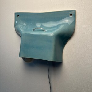 K&ouml;nnte beinhalten: Eine hellblaue Wandleuchte aus Keramik mit gl&auml;nzender Oberfl&auml;che, geschwungener organischer Form, einem kleinen silbernen Metallgriff an der Oberseite, zwei Befestigungsl&ouml;chern und einer darunter sichtbaren Gl&uuml;hbirne, verbunden mit einem d&uuml;nnen Stromkabel.