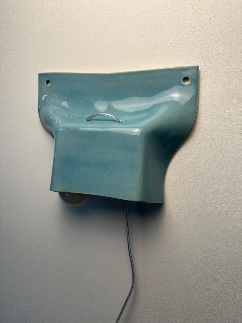 K&ouml;nnte beinhalten: Eine hellblaue Wandleuchte aus Keramik mit gl&auml;nzender Oberfl&auml;che, geschwungener organischer Form, einem kleinen silbernen Metallgriff an der Oberseite, zwei Befestigungsl&ouml;chern und einer darunter sichtbaren Gl&uuml;hbirne, verbunden mit einem d&uuml;nnen Stromkabel.