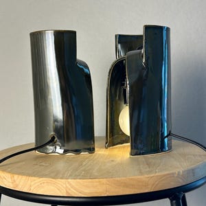 Puede incluir: Dos l&aacute;mparas de mesa de cer&aacute;mica verde oscuro con acabado brillante. Cada l&aacute;mpara tiene una estructura cil&iacute;ndrica curva con un lado abierto que revela una bombilla blanca en su interior. Un cable negro sale de la base de cada una.