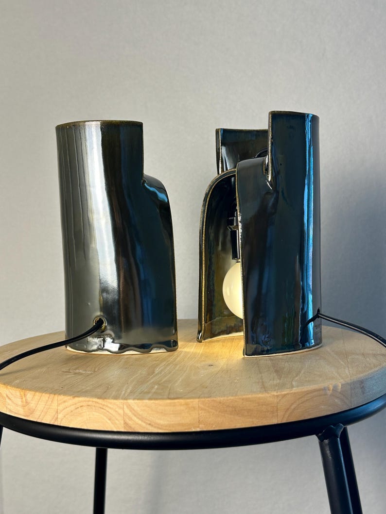 Puede incluir: Dos l&aacute;mparas de mesa de cer&aacute;mica verde oscuro con acabado brillante. Cada l&aacute;mpara tiene una estructura cil&iacute;ndrica curva con un lado abierto que revela una bombilla blanca en su interior. Un cable negro sale de la base de cada una.