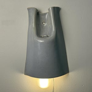 K&ouml;nnte beinhalten: Eine Wandleuchte mit einem glatten, hellgrauen Keramikschirm in einer ausgestellten, glockenf&ouml;rmigen Form. Die Leuchte verf&uuml;gt &uuml;ber ein U-f&ouml;rmiges Oberteil mit einem sichtbaren Montageloch und einer einzelnen leuchtenden Gl&uuml;hbirne unten.