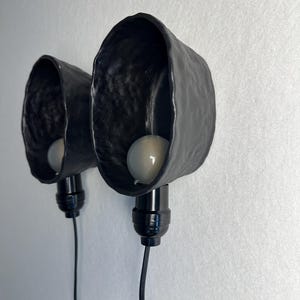 K&ouml;nnte beinhalten: Zwei Wandleuchten mit mattschwarzen, organisch geformten und texturierten Keramikschirmen. Jede Leuchte besteht aus einer schwarzen Fassung, einem schwarzen Stromkabel und einer runden wei&szlig;en Gl&uuml;hbirne im Inneren des tiefen Keramikgeh&auml;uses.