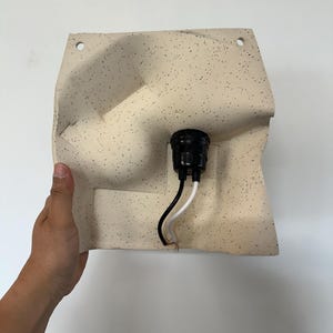 May include: A wall-mounted light fixture featuring a cream-colored, speckled ceramic shade with an irregular, molded shape. The fixture includes a central black socket with one black and one white electrical wire extending from the bottom.