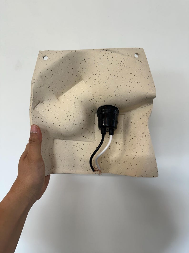 May include: A wall-mounted light fixture featuring a cream-colored, speckled ceramic shade with an irregular, molded shape. The fixture includes a central black socket with one black and one white electrical wire extending from the bottom.