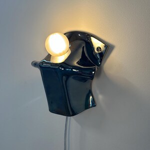 May include: A wall-mounted lamp featuring a glossy, dark, reflective, organic-shaped ceramic or metal base with a single, glowing, spherical light bulb positioned on top. A thin, light-colored power cord hangs vertically from the bottom of the fixture.