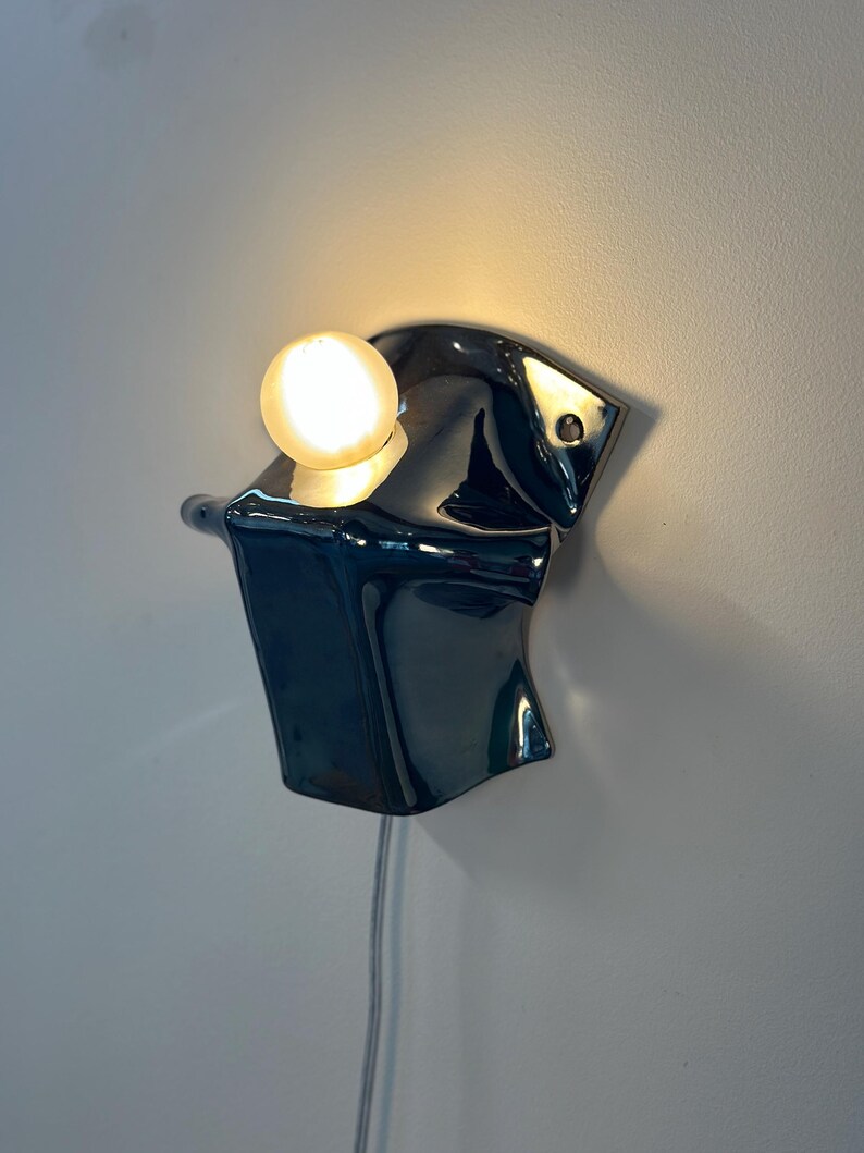 May include: A wall-mounted lamp featuring a glossy, dark, reflective, organic-shaped ceramic or metal base with a single, glowing, spherical light bulb positioned on top. A thin, light-colored power cord hangs vertically from the bottom of the fixture.