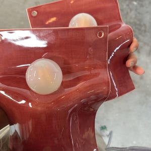 May include: Two wall-mounted ceramic sconces with a glossy, dusty rose finish. Each piece features a curved, wave-like form with a central circular opening holding a frosted white glass globe light bulb. Small holes are visible at the top corners.